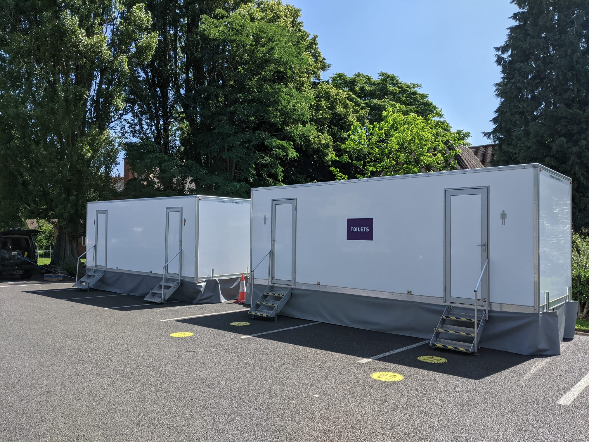 Two white luxury toilet trailers with toilets signage parked at a summer event surrounded by trees